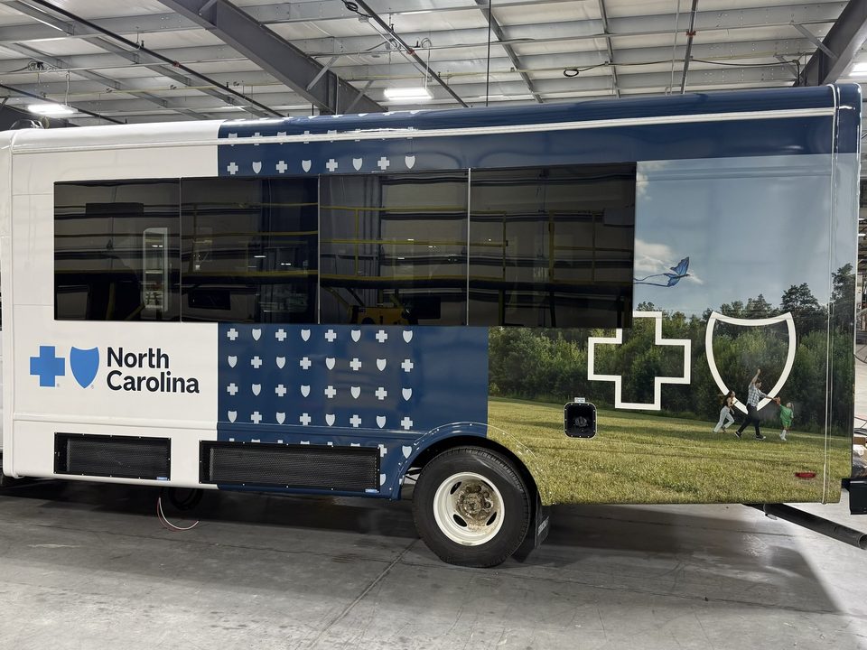 Mobile outreach bus wrapped navy with a cross-and-shield pattern and a photo panel, driver side, in the plant