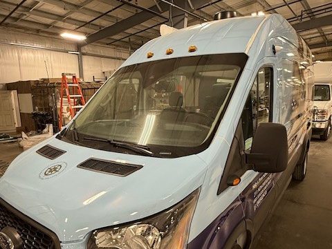 Light-blue and navy Ford Transit high-roof van wrapped for The Encompassing Center, front three-quarter, in the HLE plant