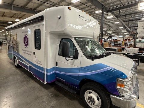 Mobile food pantry on a Ford E-Series cutaway, white with a blue and purple Catholic Charities wrap, front three-quarter, in the HLE plant