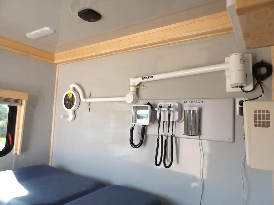 Exam room inside a mobile medical unit: wall-mounted diagnostic set, exam table, cabinetry