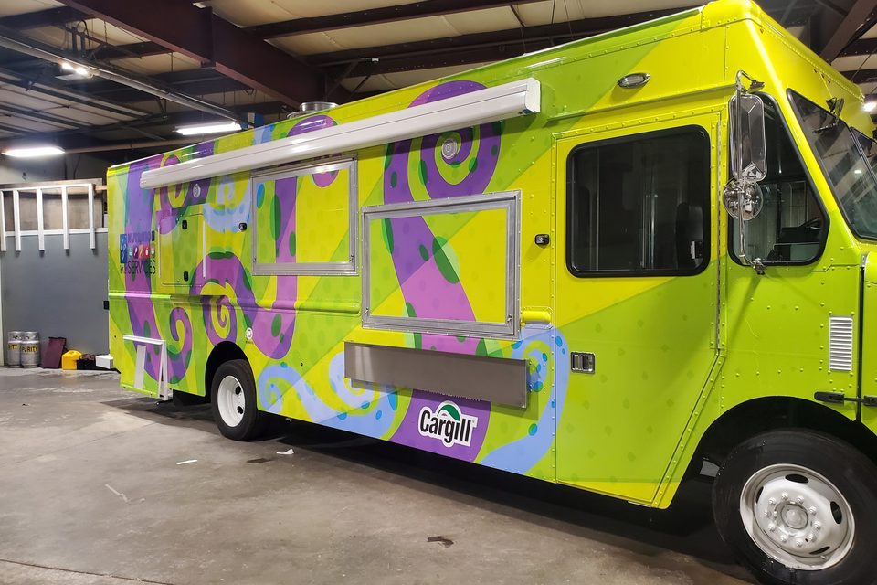 Mobile kitchen wrapped green and purple for the Saint Paul Public Schools school-lunch program, front three-quarter, in the plant
