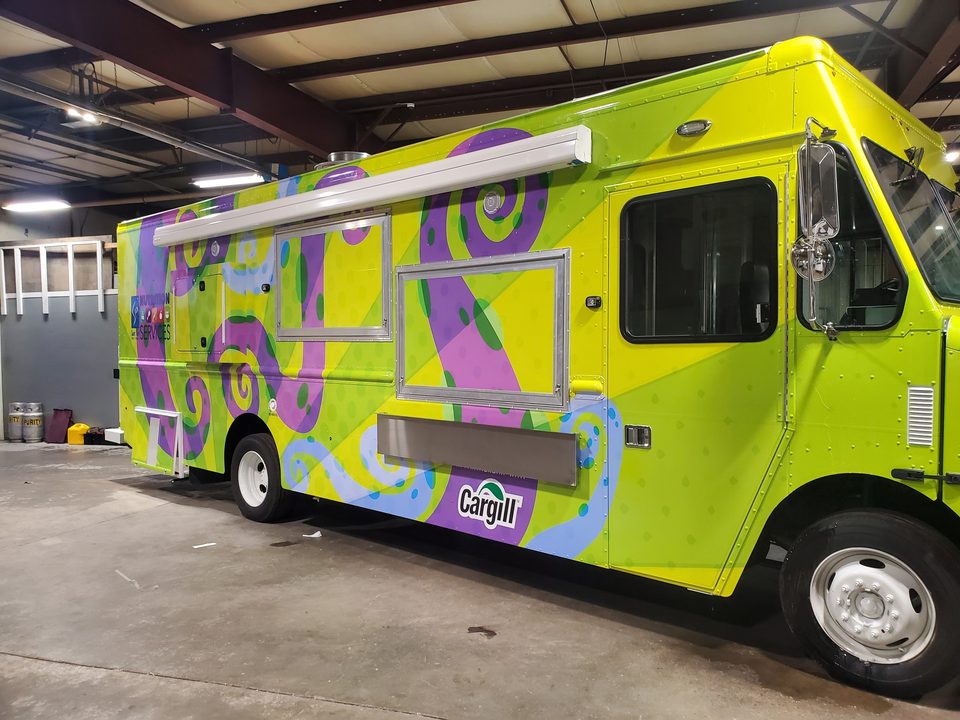 Mobile kitchen wrapped green and purple for the Saint Paul Public Schools school-lunch program, front three-quarter, in the plant