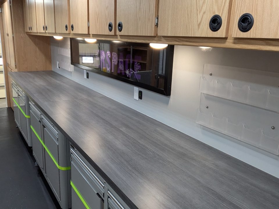 Makerspace interior: long work counter, grey cabinets with green accents, oak overheads