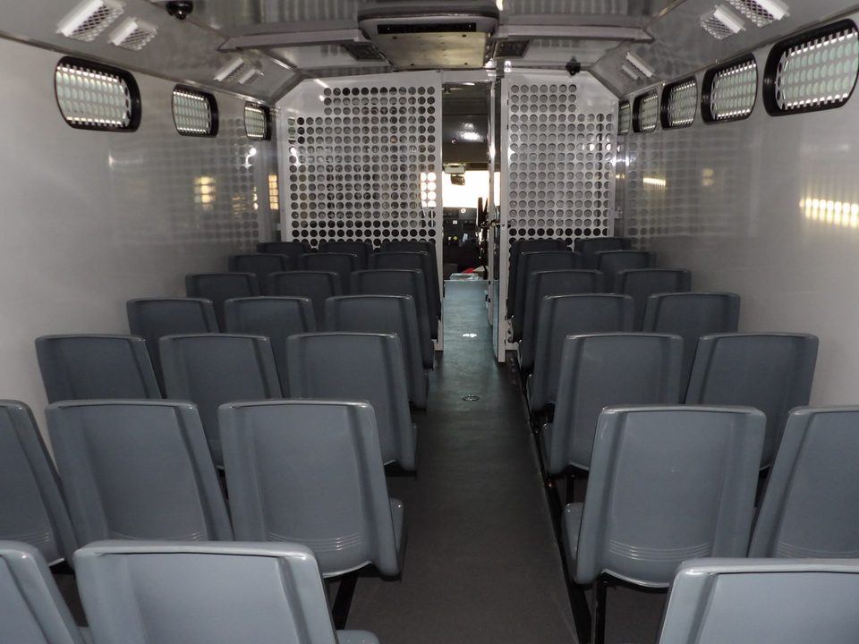Prisoner transport interior looking aft: rows of grey molded seats both sides of the aisle, slot windows, perforated-steel partition at the rear