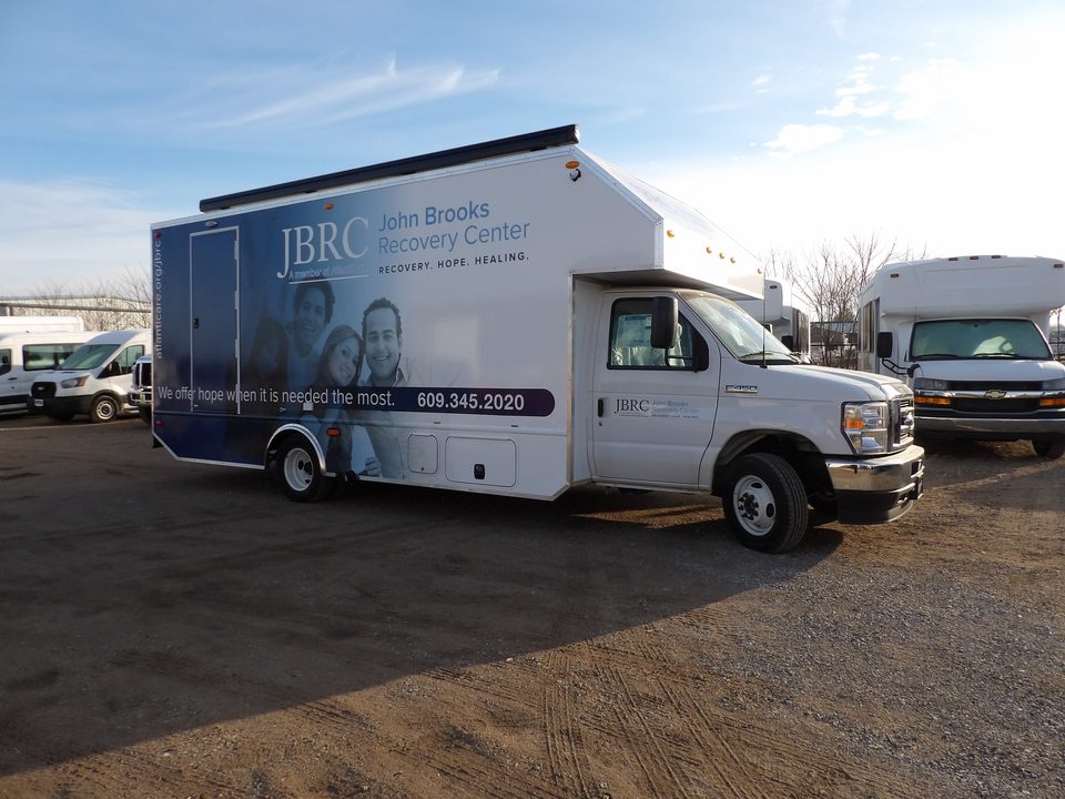 Mobile recovery unit on a Ford E-450 cutaway, blue and white John Brooks Recovery Center wrap, front three-quarter, blue sky