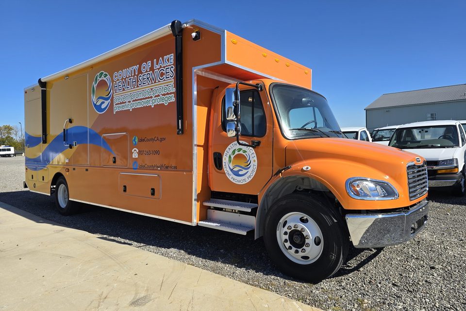 Mobile health clinic on a Freightliner M2, orange and blue County of Lake Health Services wrap, front three-quarter, blue sky