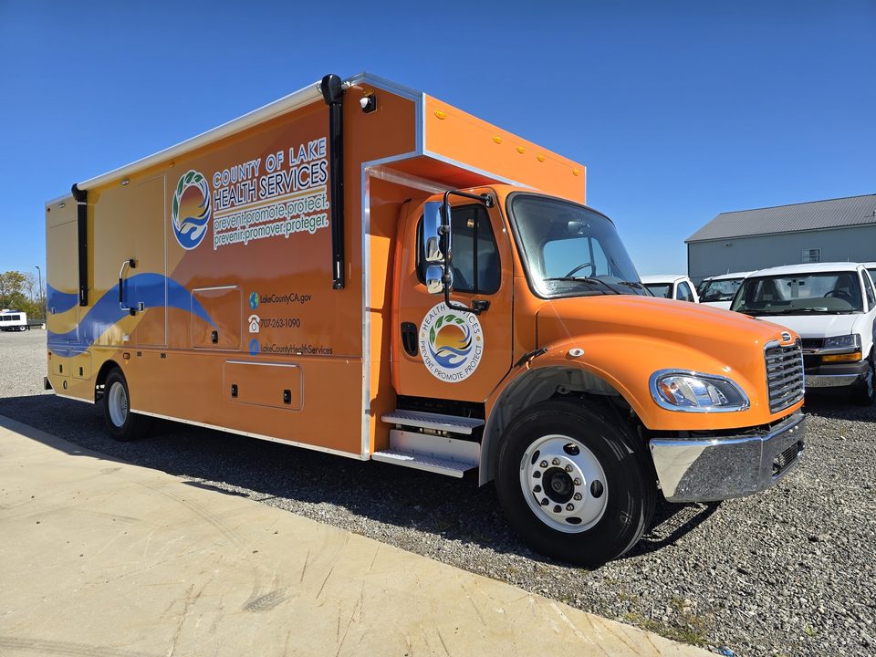 Mobile health clinic on a Freightliner M2, orange and blue County of Lake Health Services wrap, front three-quarter, blue sky