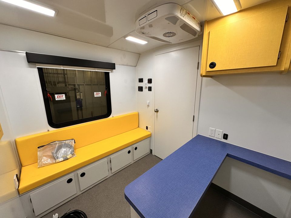 Outreach unit interior: yellow bench seating with storage, L-shaped blue counter, overhead cabinet