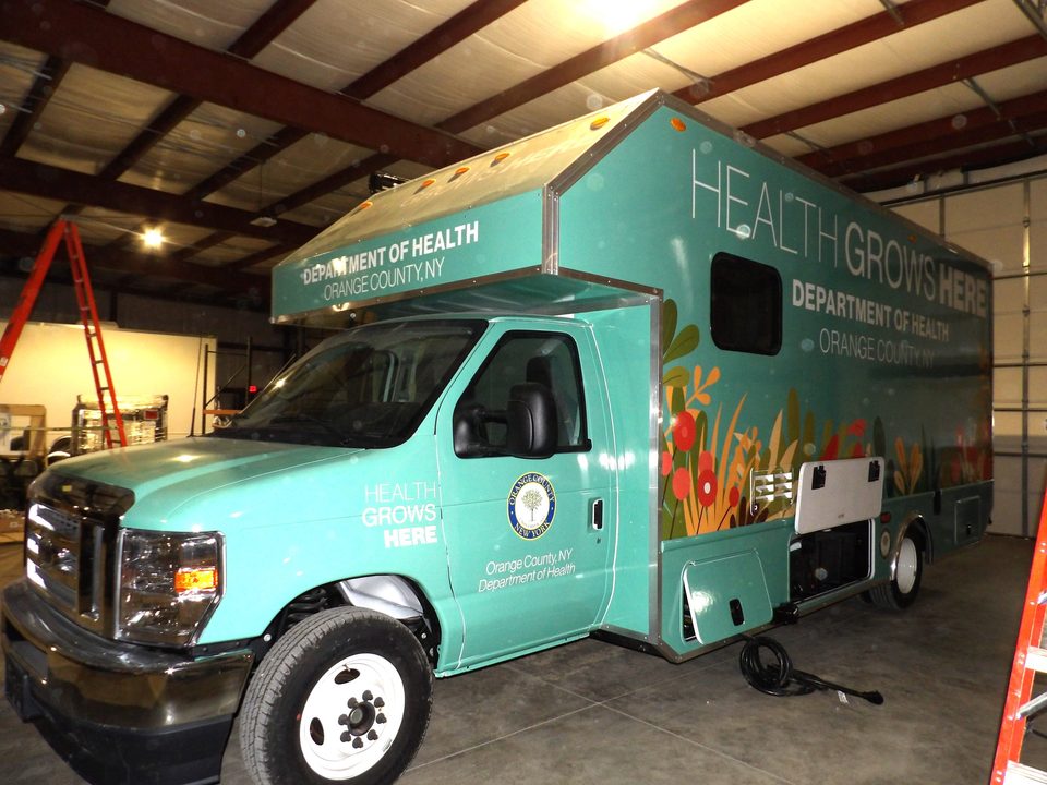 Mobile health unit on a Ford E-450 cutaway, teal Health Grows Here wrap for the Orange County Department of Health, front three-quarter, in the HLE plant