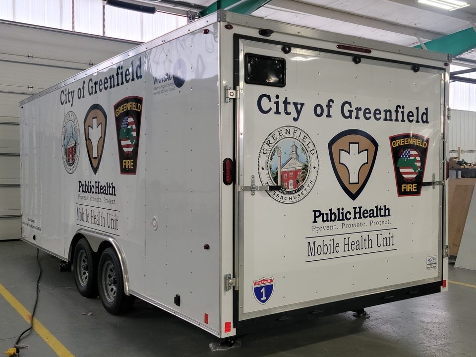 White bumper-pull trailer lettered City of Greenfield Public Health Mobile Health Unit, rear three-quarter, in the HLE plant