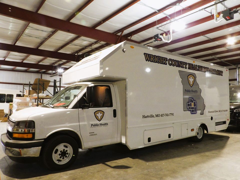 Mobile public-health clinic on a Chevrolet 4500 cutaway, Wright County Health Department wrap, front three-quarter, in the HLE plant